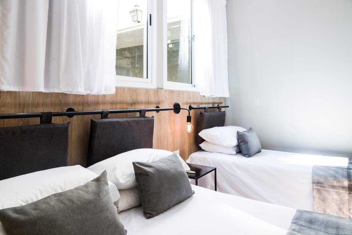 Ein Hotelzimmer mit zwei Betten, einem Holzkopfende, weißer Bettwäsche und Blick ins Freie durch ein Fenster mit weißen Vorhängen.
