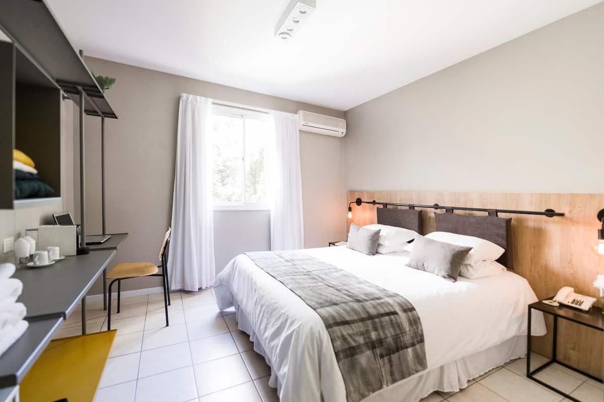 Ein modernes Hotelzimmer mit Bett, Nachttisch, Sessel, Schreibtisch und einem Fenster mit Vorhängen.