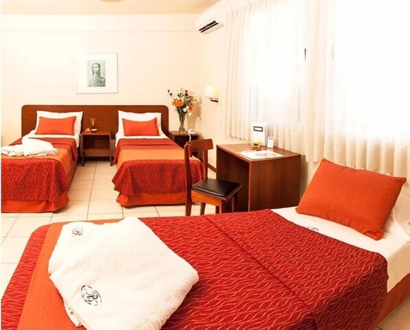 Habitación de hotel con dos camas, cada una con ropa de cama y almohadas rojas, un escritorio pequeño con lámpara y una ventana con cortinas.