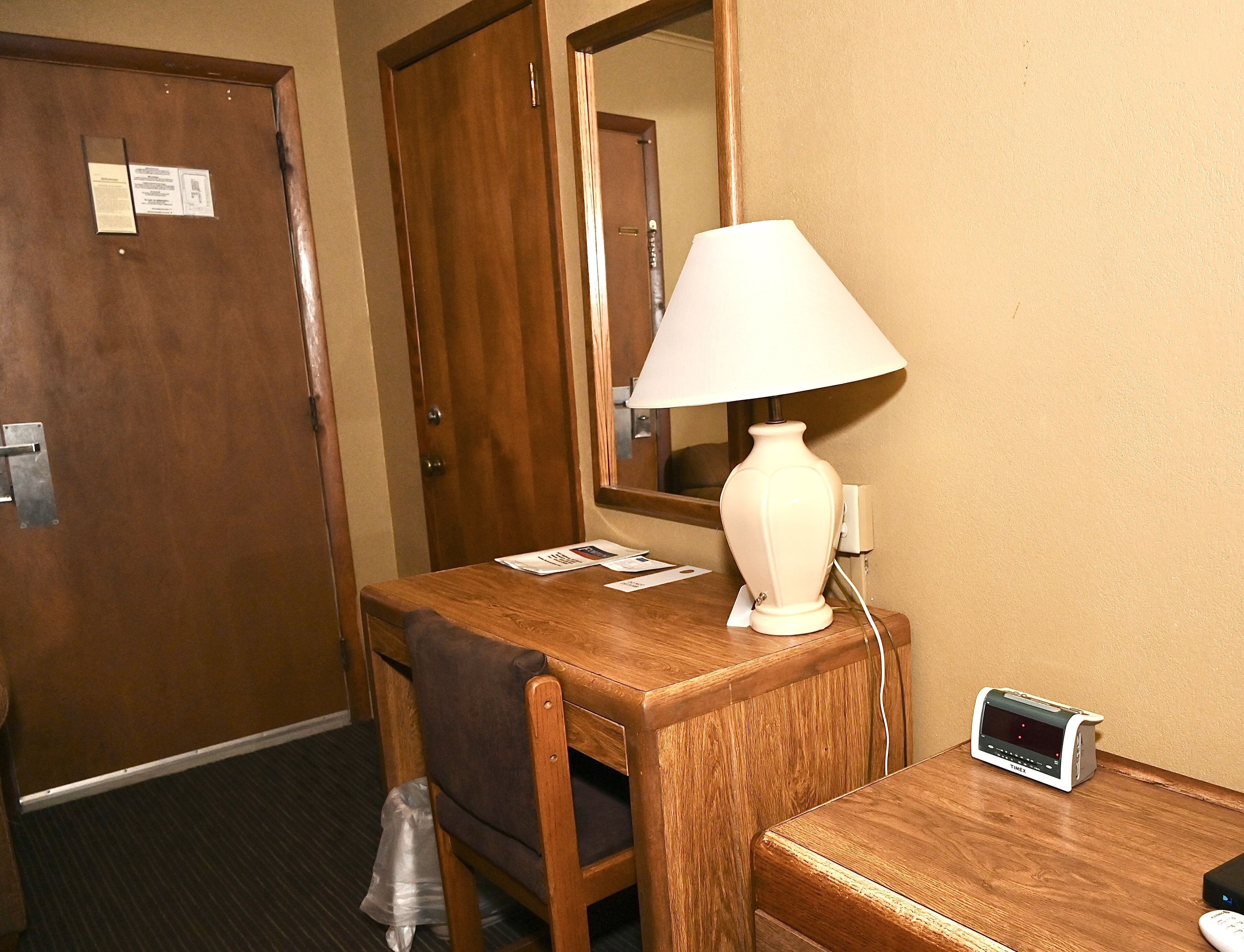 A hotel room with a wooden desk, a lamp, a clock, and a trash bag on the floor.