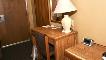 A hotel room with a wooden desk, a lamp, a clock, and a trash bag on the floor.