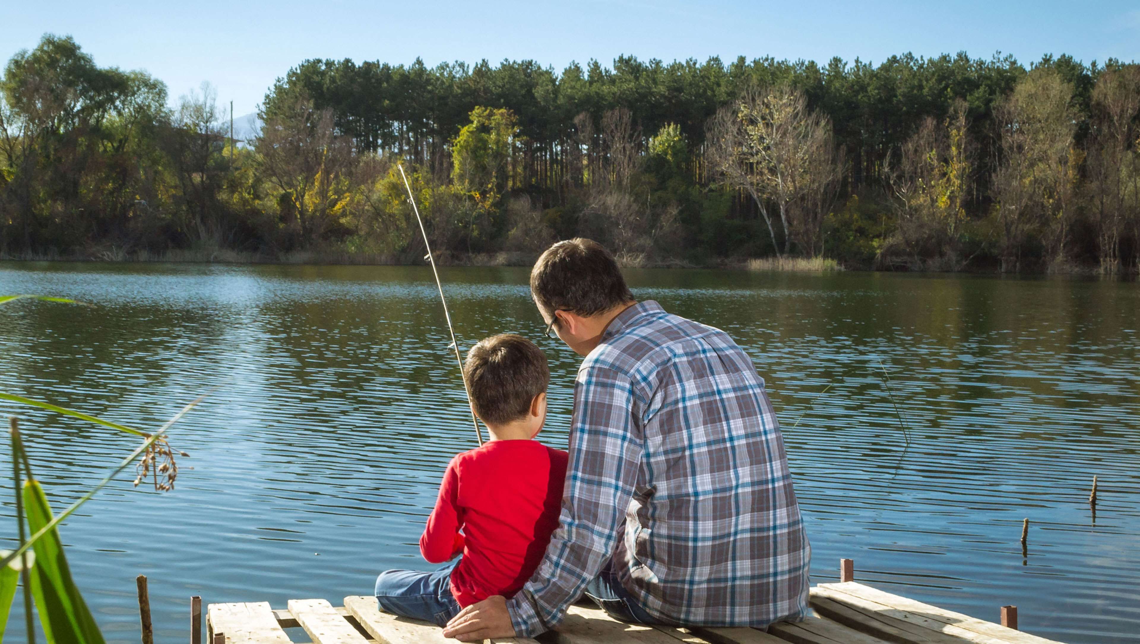 Un hombre y un niño sentados en un muelle de madera junto a un lago tranquilo, rodeados de árboles.