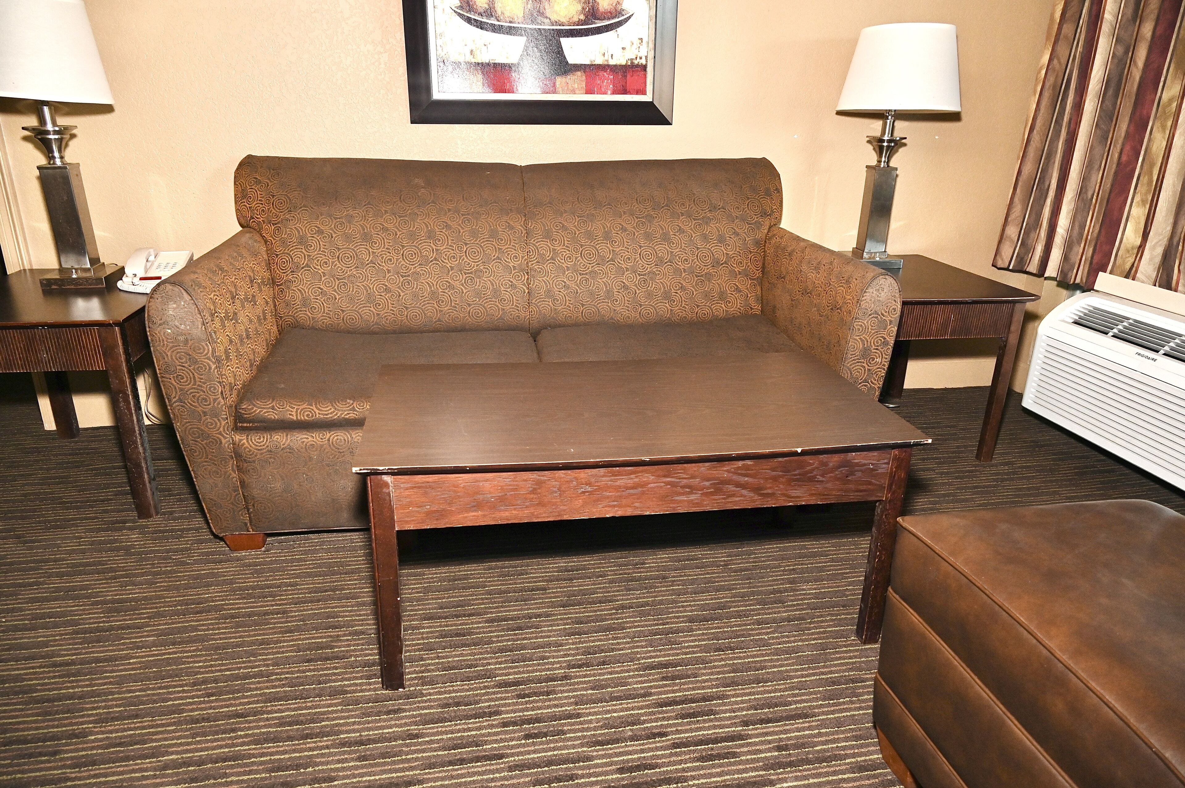 Quarto de hotel com um sofá marrom, uma mesa de centro de madeira, uma pequena mesa lateral com uma luminária e um quadro na parede.