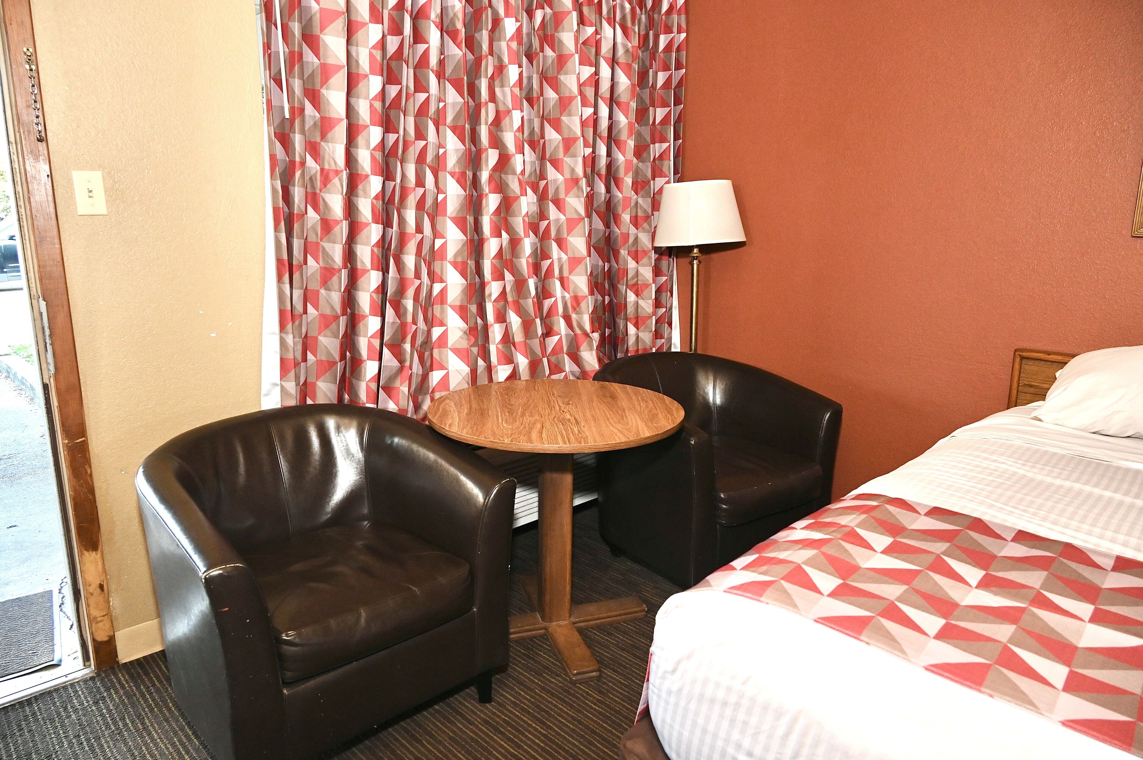 Une chambre d’hôtel avec un lit, deux fauteuils en cuir, une petite table en bois, une lampe et un rideau à motifs.