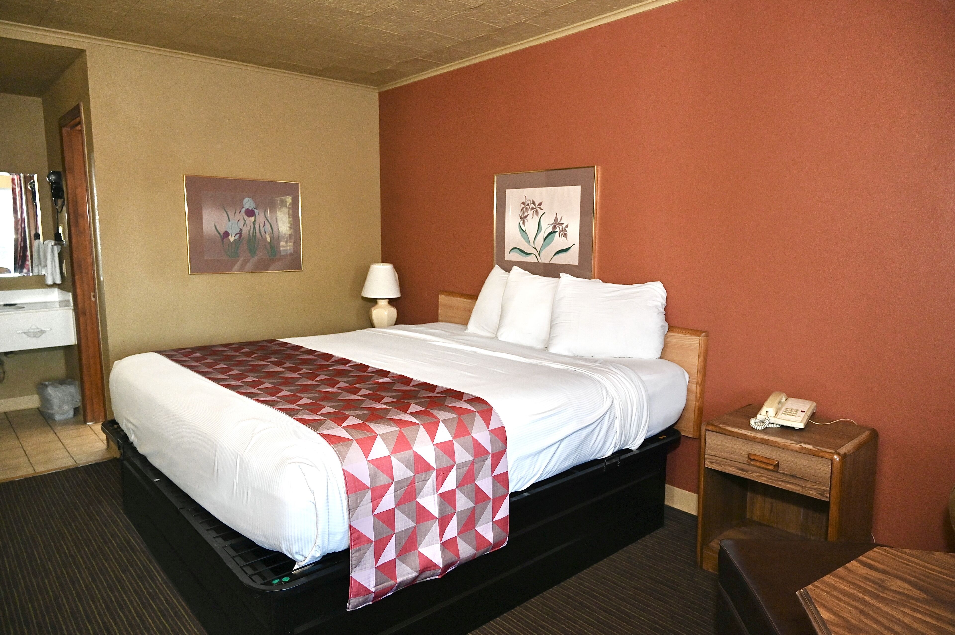 A hotel room with a bed, a nightstand with a phone, a lamp, and two framed pictures on the wall.