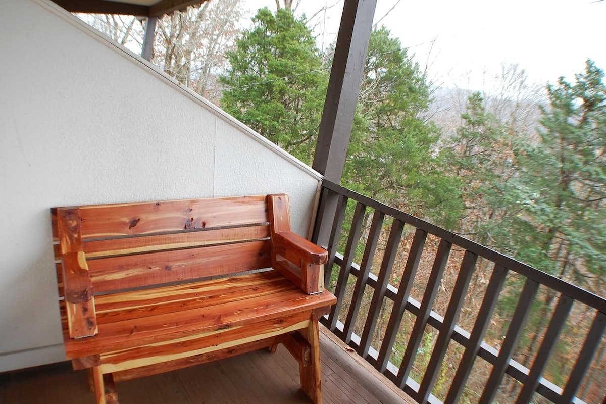 A wooden bench on a balcony with a railing, overlooking a forested area.