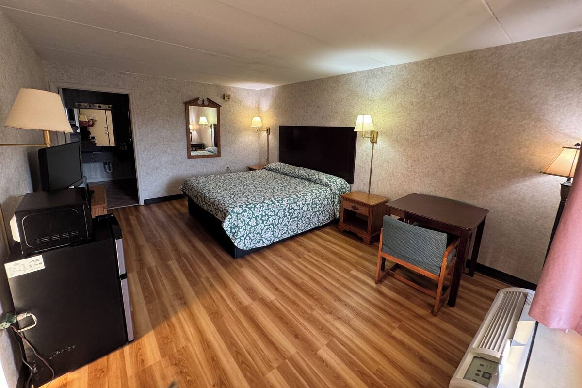 A compact hotel room with a bed, bedside table, desk, and a small kitchenette area.