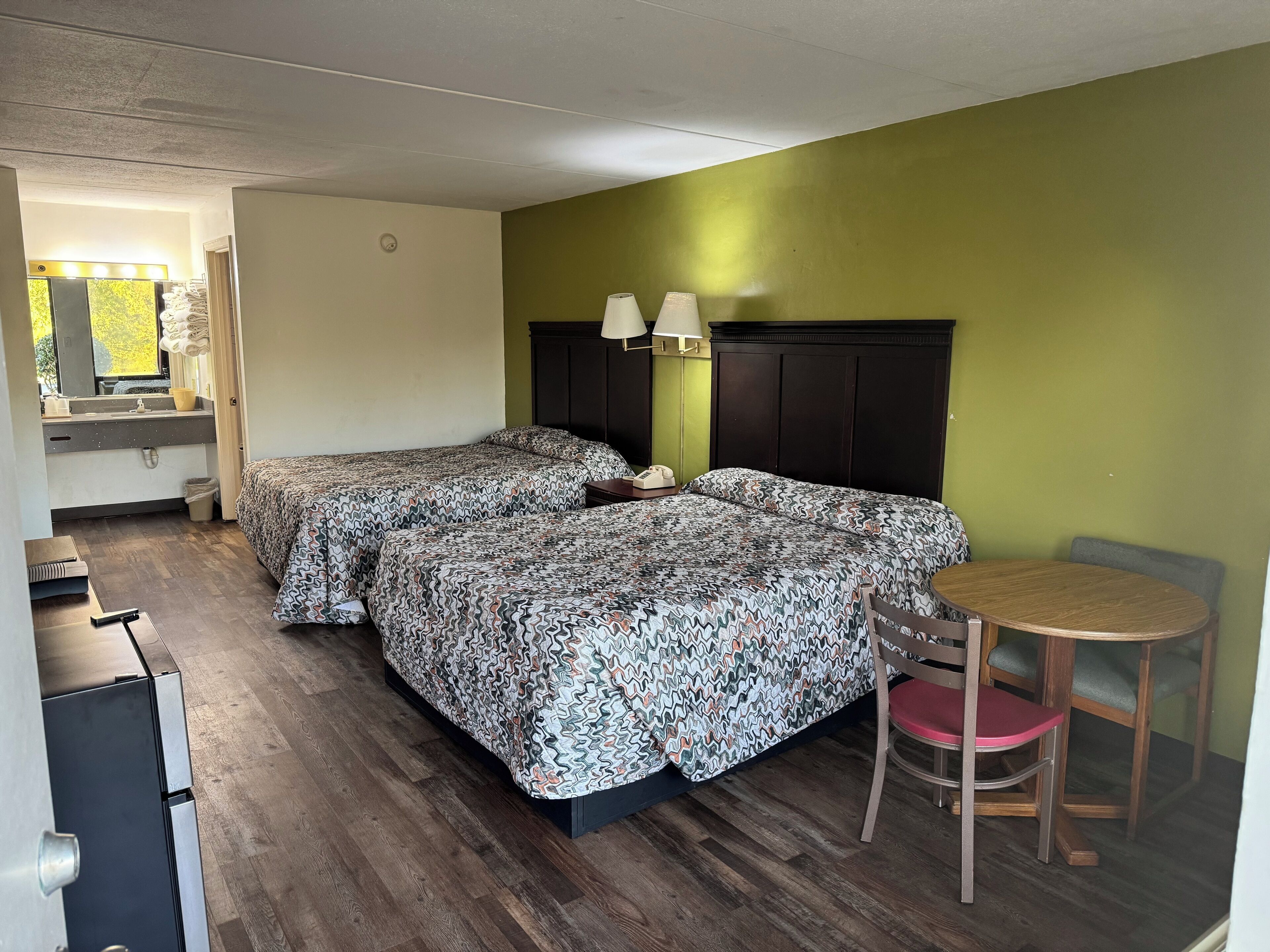 A hotel room with two beds, a small table, a chair, a desk, and a mirror with lights.
