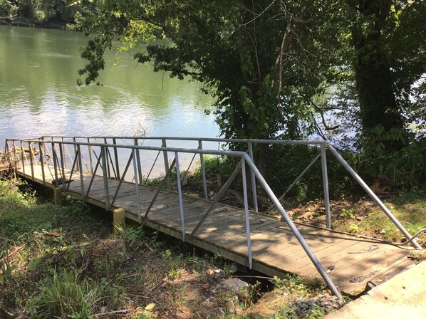 A wooden dock with metal railings extending over a body of water, surrounded by greenery. - Riverfront Lodge (Burkesville)