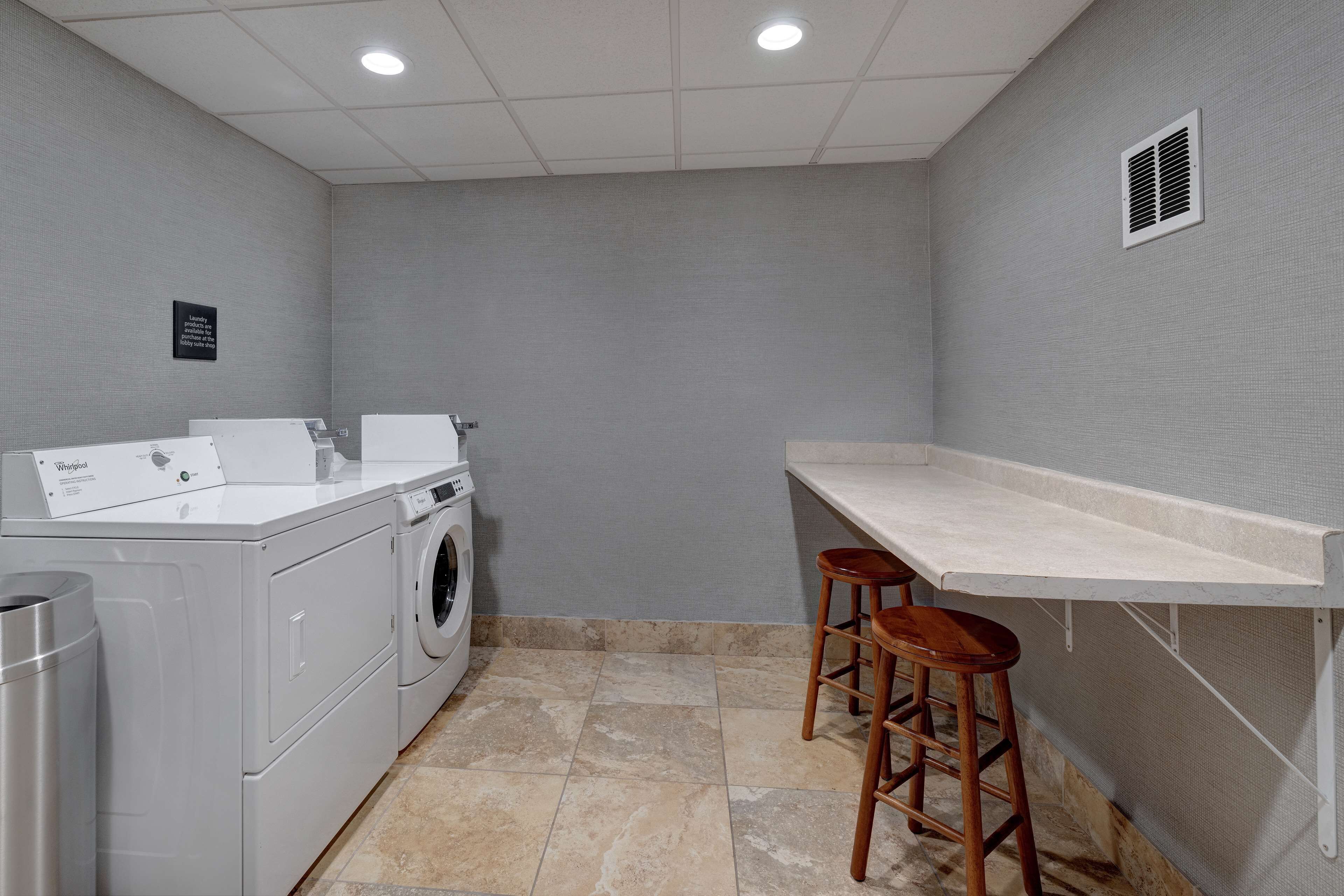 A laundry room with a front-loading washer and dryer, a trash can, and a bar stool.