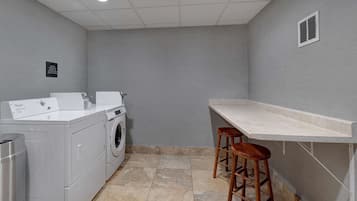 A laundry room with a front-loading washer and dryer, a trash can, and a bar stool.