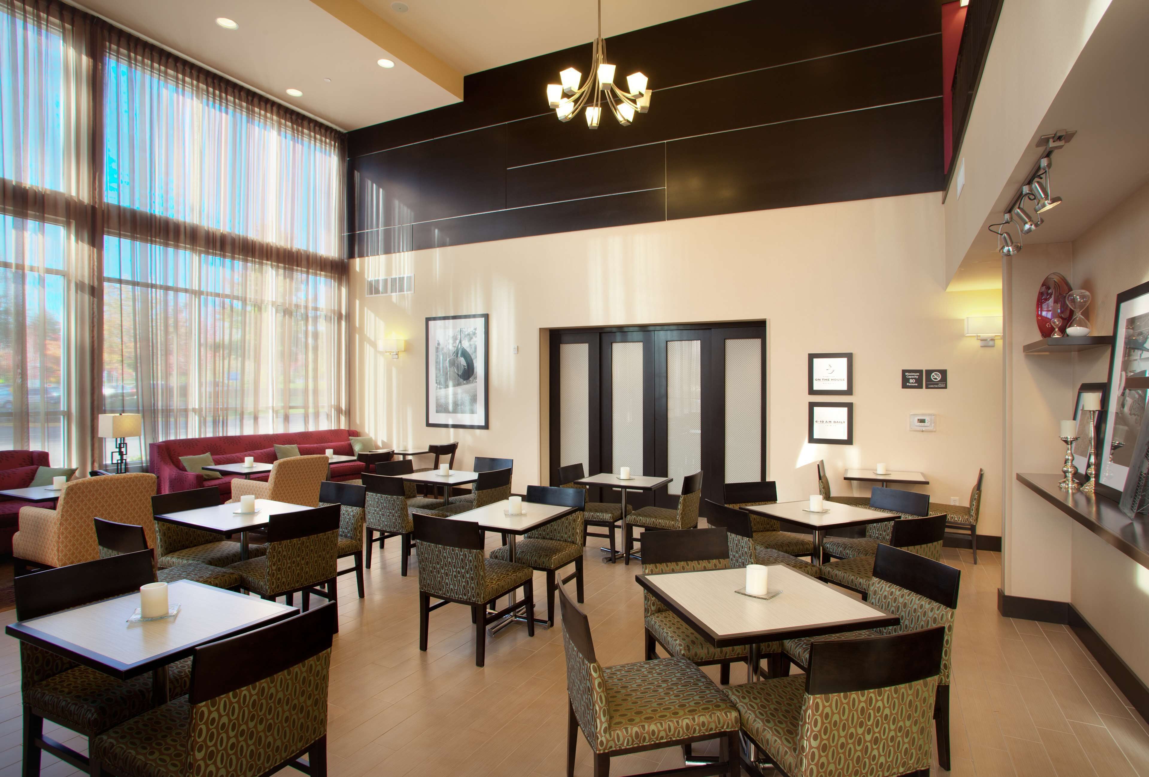 Un comedor de hotel con varias mesas y sillas, ventanas amplias con cortinas translúcidas y un candelabro moderno.