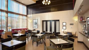 Un comedor de hotel con varias mesas y sillas, ventanas amplias con cortinas translúcidas y un candelabro moderno.