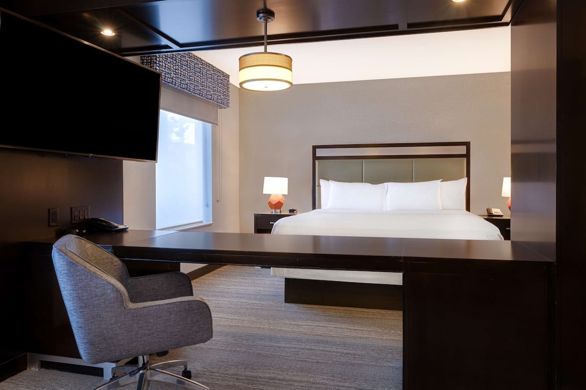 A hotel room with a large bed, a desk with a chair, a flat-screen TV, and a lamp.