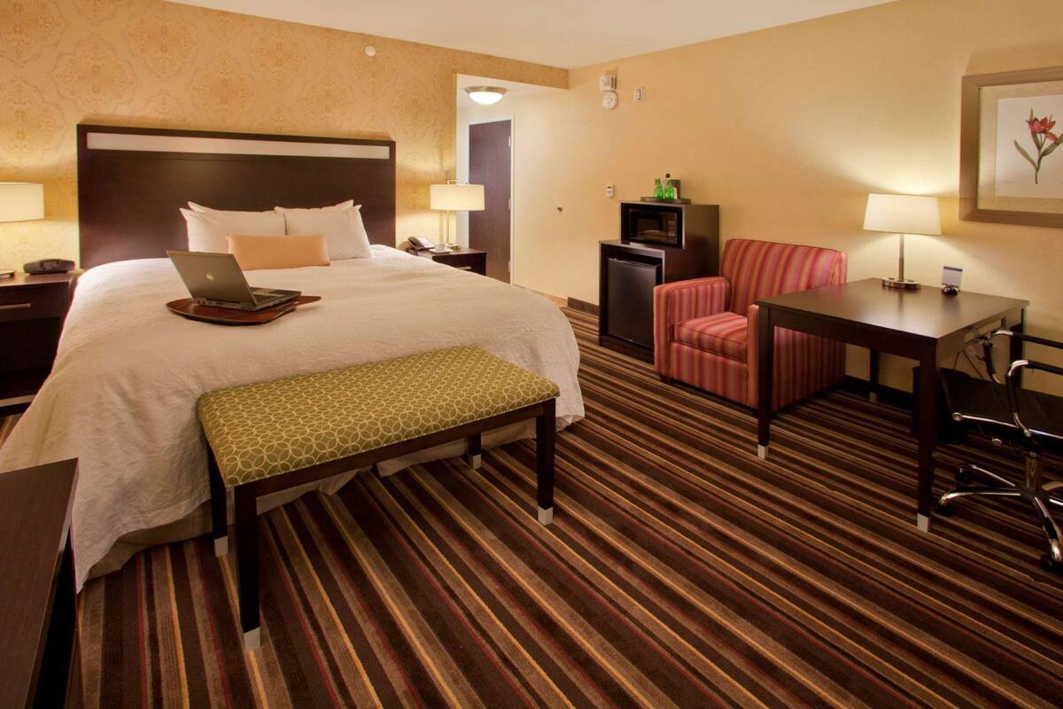 A hotel room with a large bed, a desk with a chair, a small table with a chair, a bench, and a TV on a cabinet.