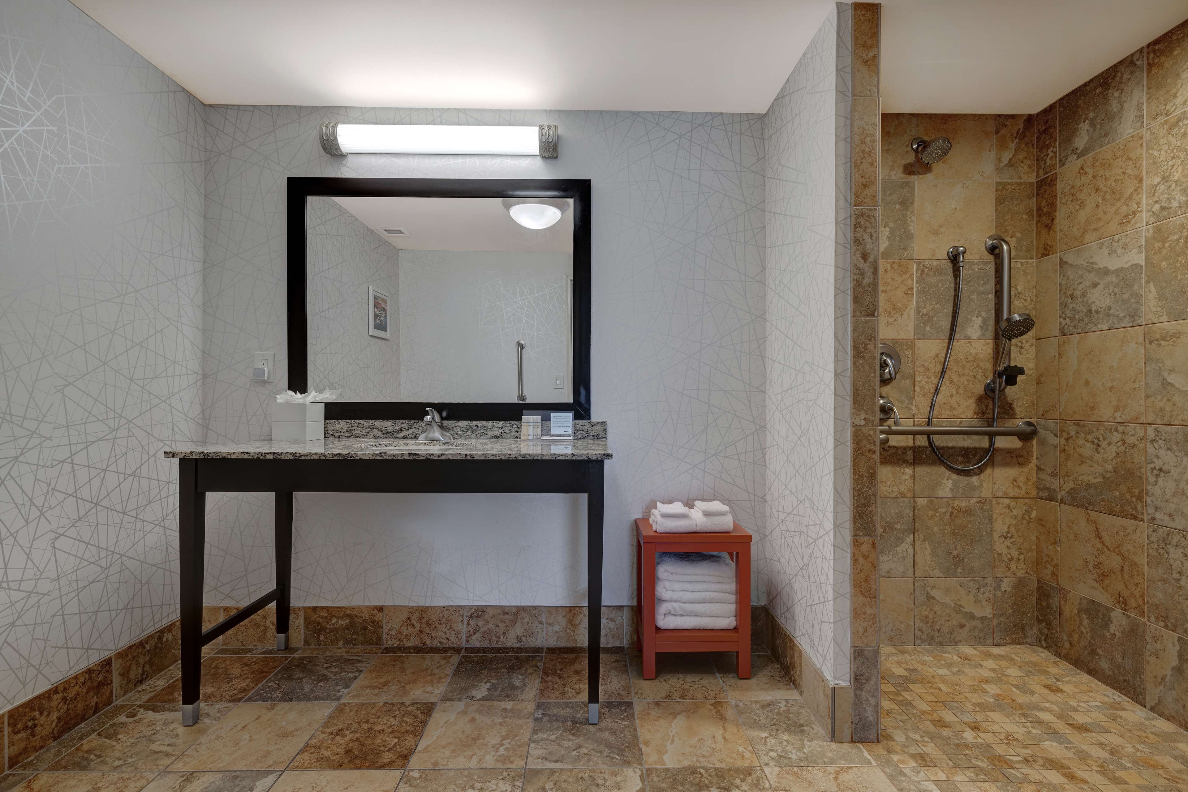 A bathroom with a granite countertop sink, a mirror, a small stool with towels, and a shower area with a handheld showerhead.