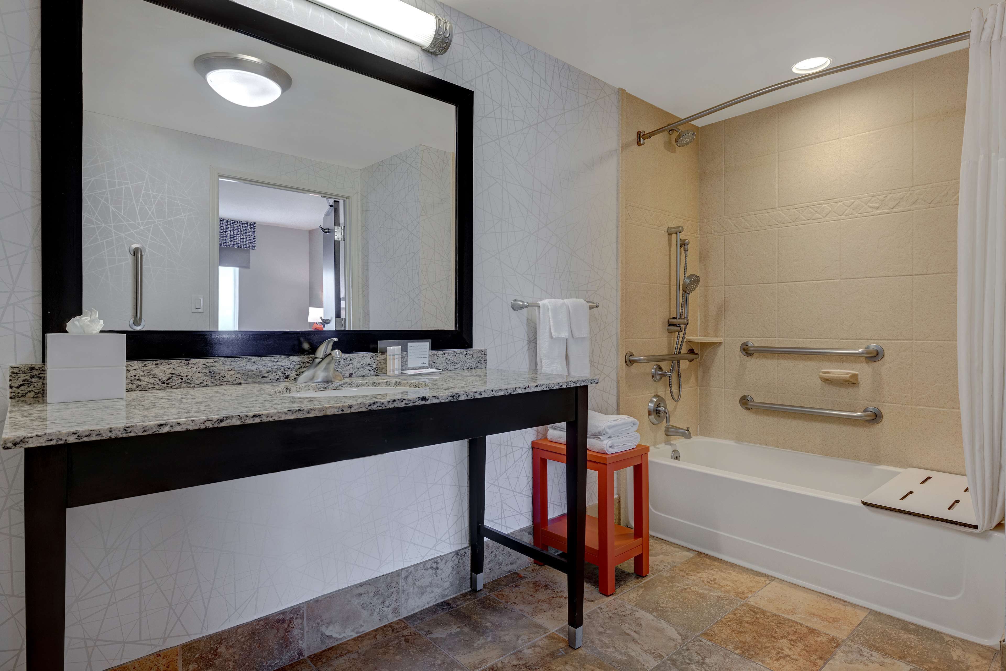A modern bathroom with a large mirror, a granite countertop, a shower with grab bars, and a small stool.