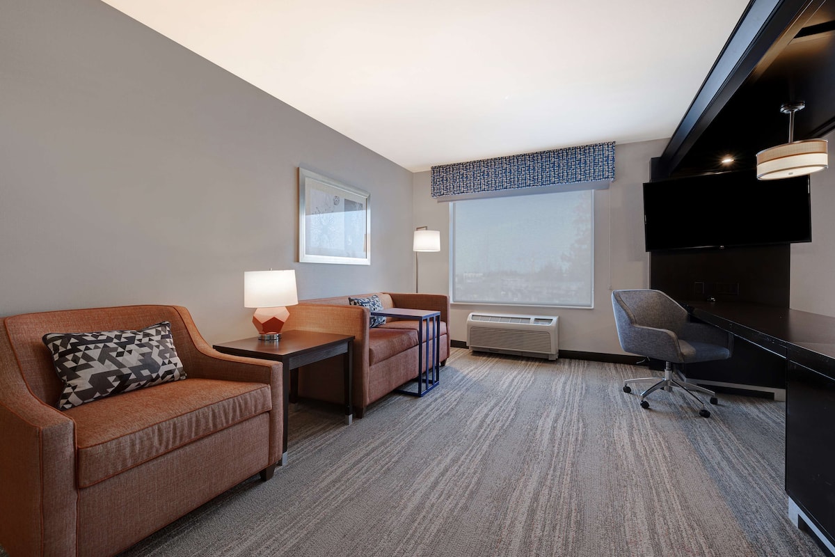 A hotel room with a sofa, a small table, a chair, a TV, and a large window.