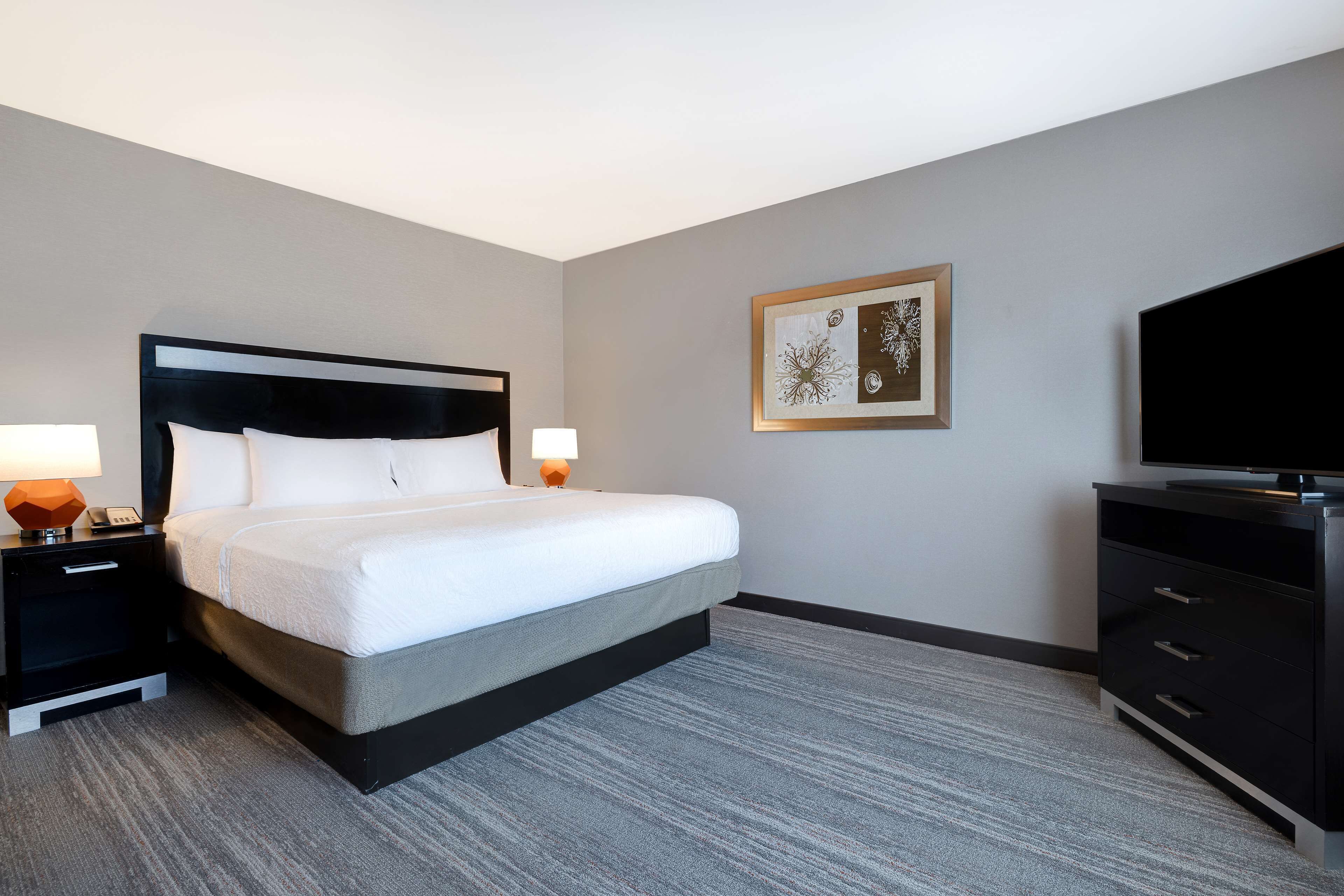 A hotel room with a bed, bedside tables, a TV, and a framed artwork on the wall.