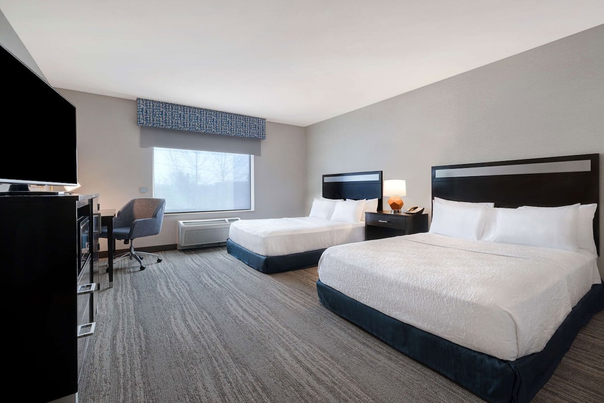 A hotel room with two beds, a desk, a chair, a TV, and a large window.