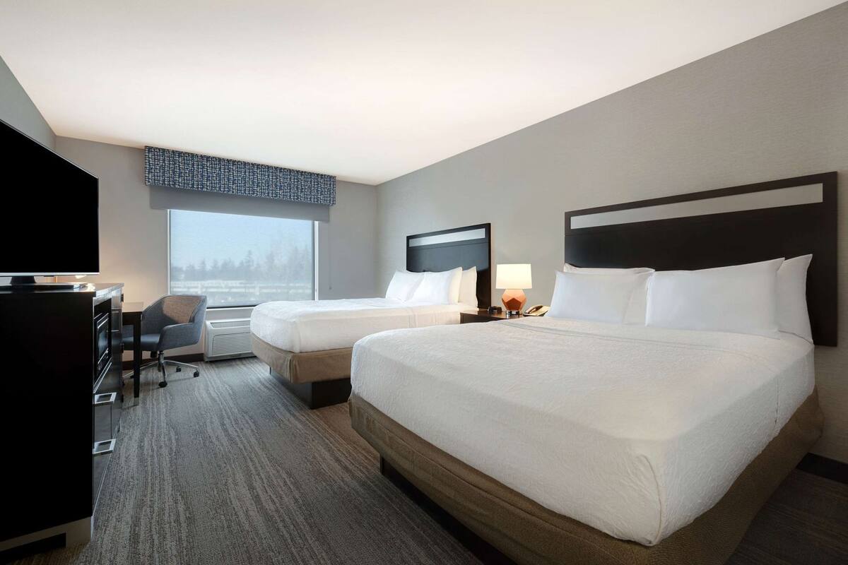 A hotel room with two beds, a desk, a chair, a TV, and a window with a view.