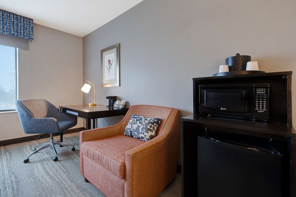 A hotel room with a grey wall, a framed picture, a grey office chair, a small orange sofa, a black microwave, and a black refrigerator.