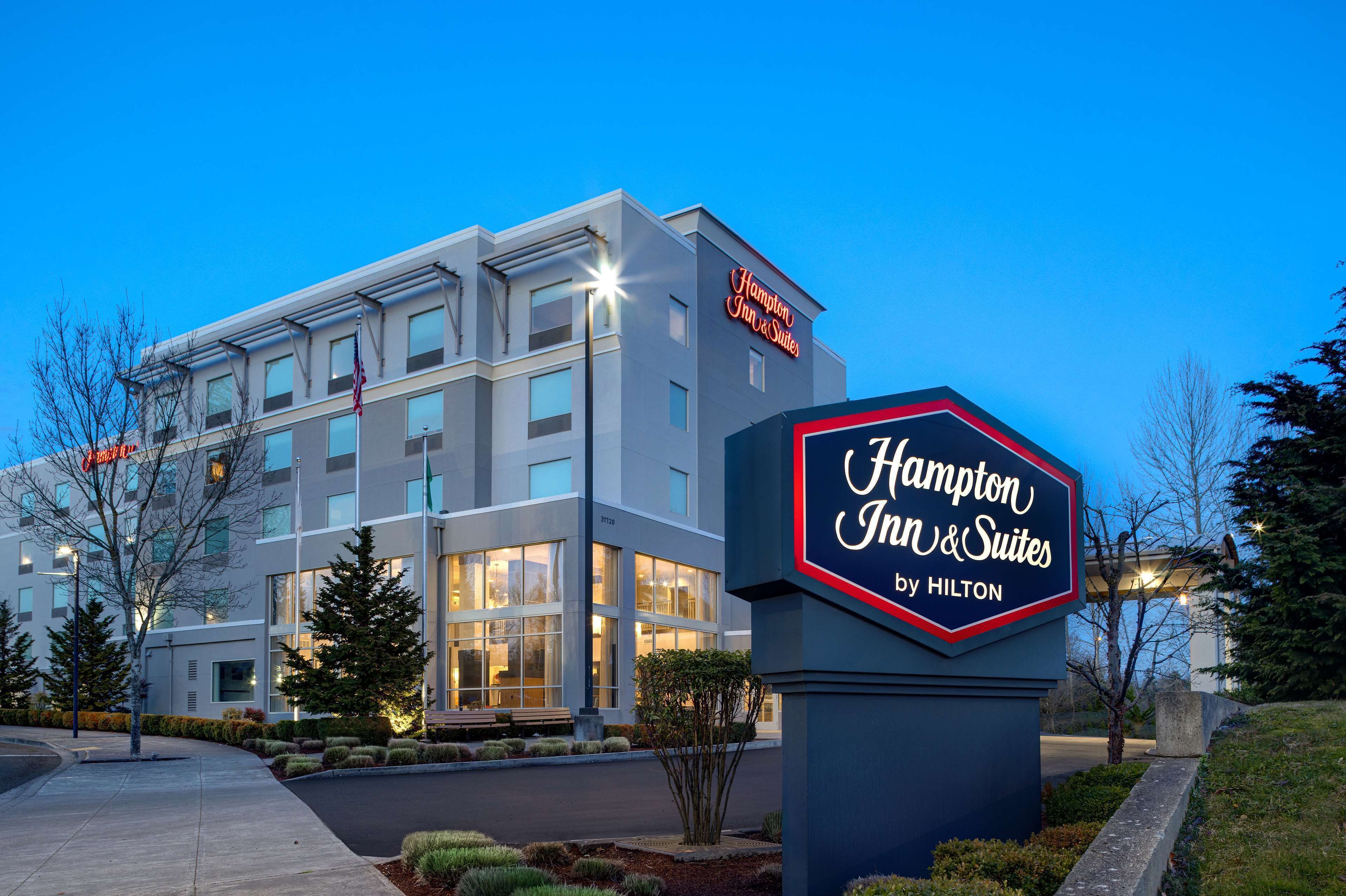 Un bâtiment de la chaîne Hampton Inn & Suites by Hilton, avec une enseigne bien visible, de grandes fenêtres et une entrée bien éclairée.