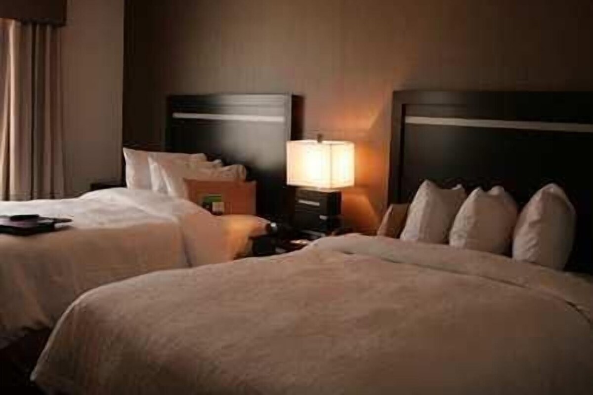 Una habitación de hotel con dos camas, una mesita de noche, una lámpara y un ventanal con cortinas.
