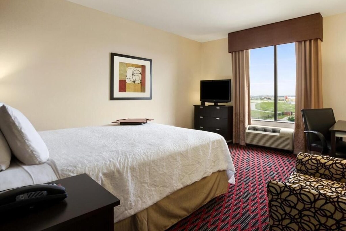 Habitación de hotel con cama, televisión, escritorio con silla, ventana con vistas y un cuadro en la pared.