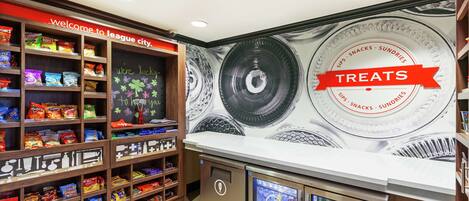 Un espace dédié à la vente de collations, avec des étagères remplies de différents types de produits et un panneau indiquant : « Bienvenue à League City. »