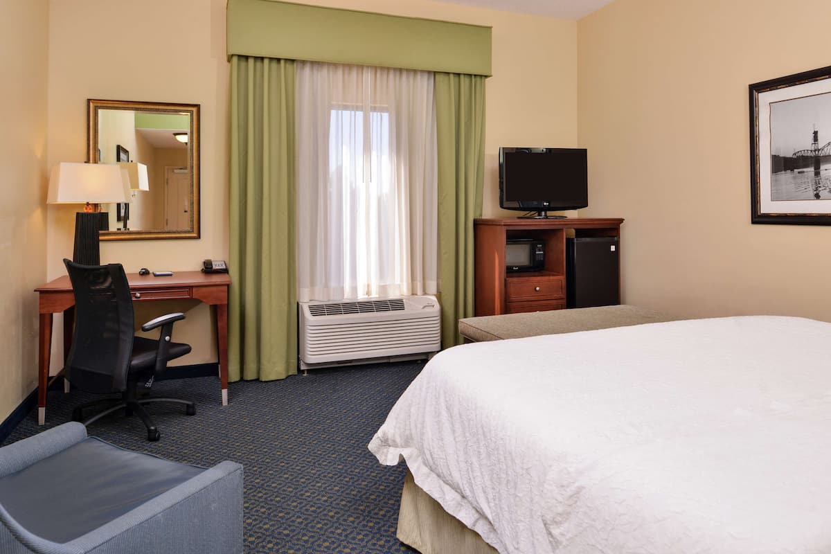 A hotel room with a bed, desk, chair, TV, and air conditioning unit.