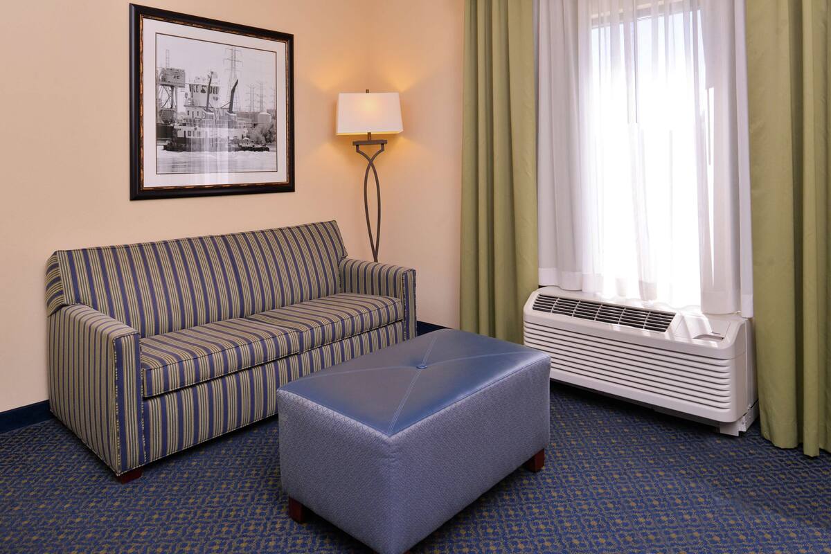 A hotel room with a striped sofa, a tufted ottoman, a framed black and white photo on the wall, a floor lamp, and a window with curtains.