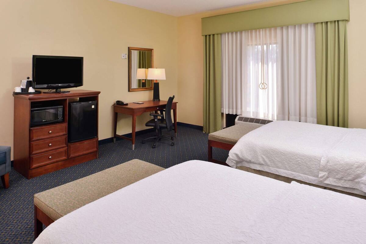 A hotel room with two beds, a desk with a chair, a television, a microwave, and a lamp.