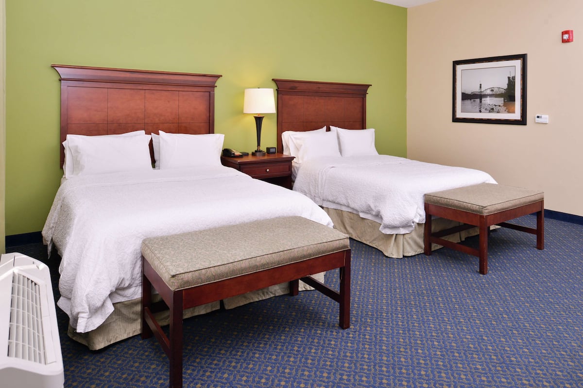A hotel room with two beds, a bench, a bedside table with a lamp, and a framed picture on the wall.