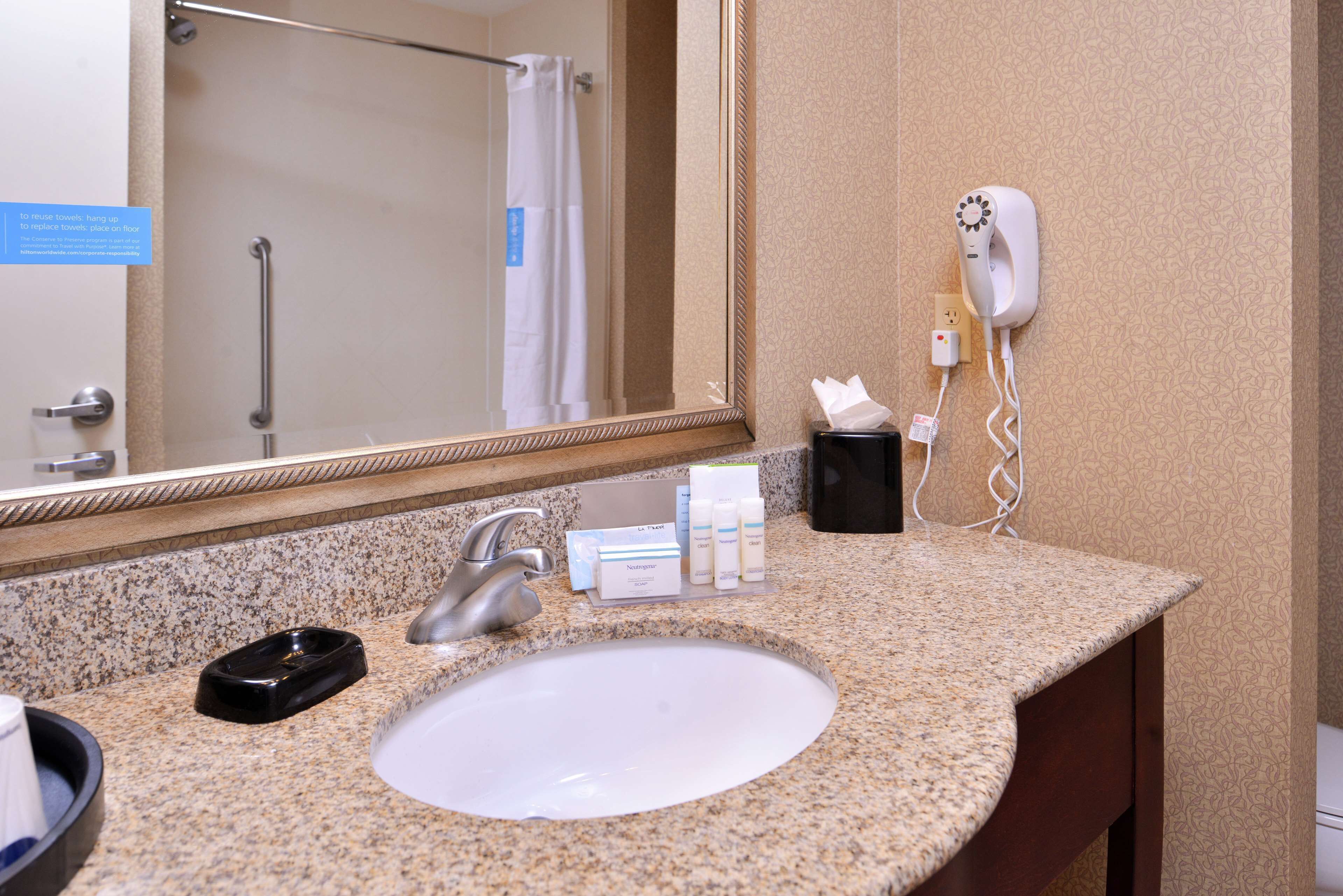 A bathroom with a granite countertop, a sink, a mirror, a towel rack, a hairdryer, and toiletries.