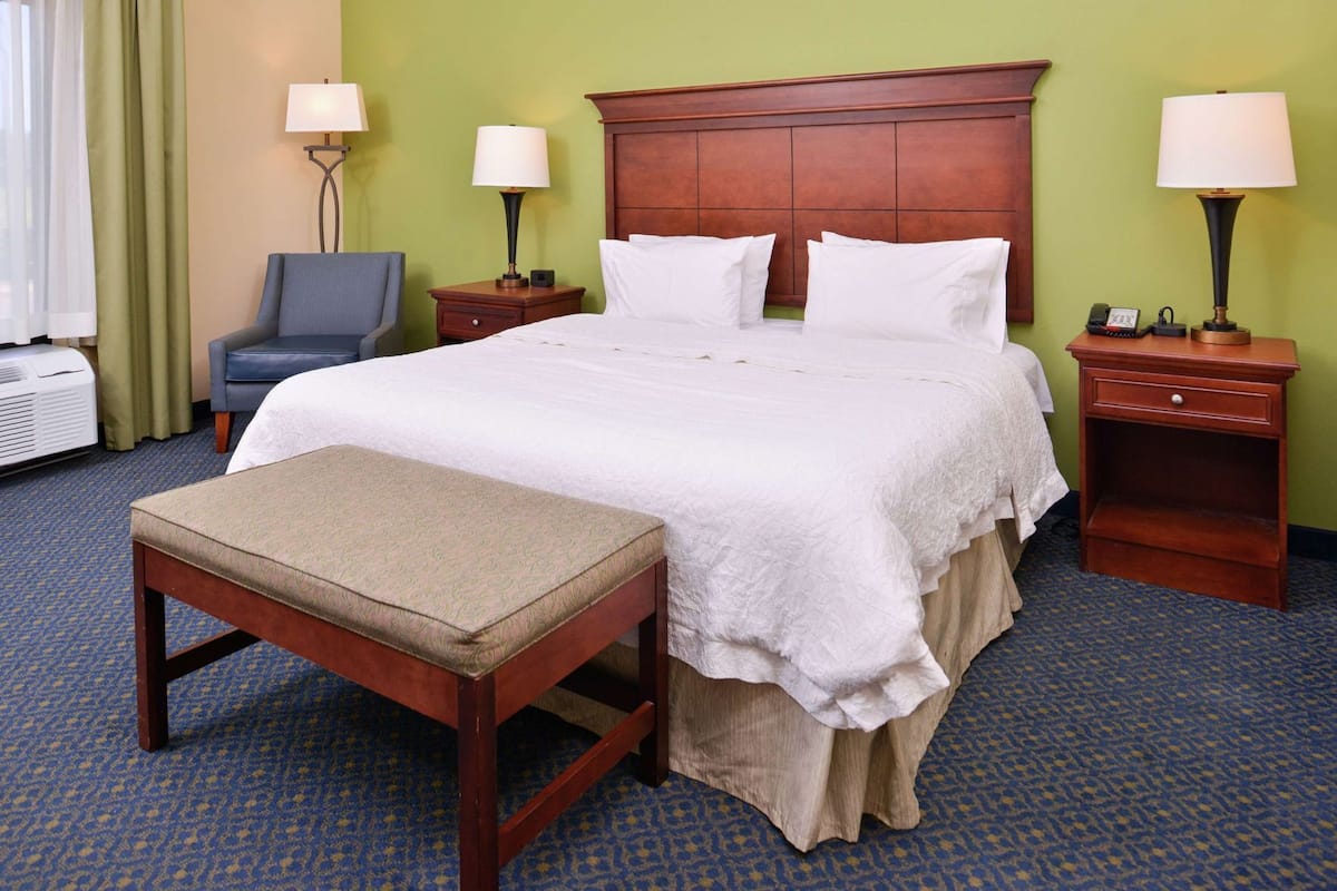 A hotel room with a large bed, a bedside table, a chair, and a desk with a lamp.