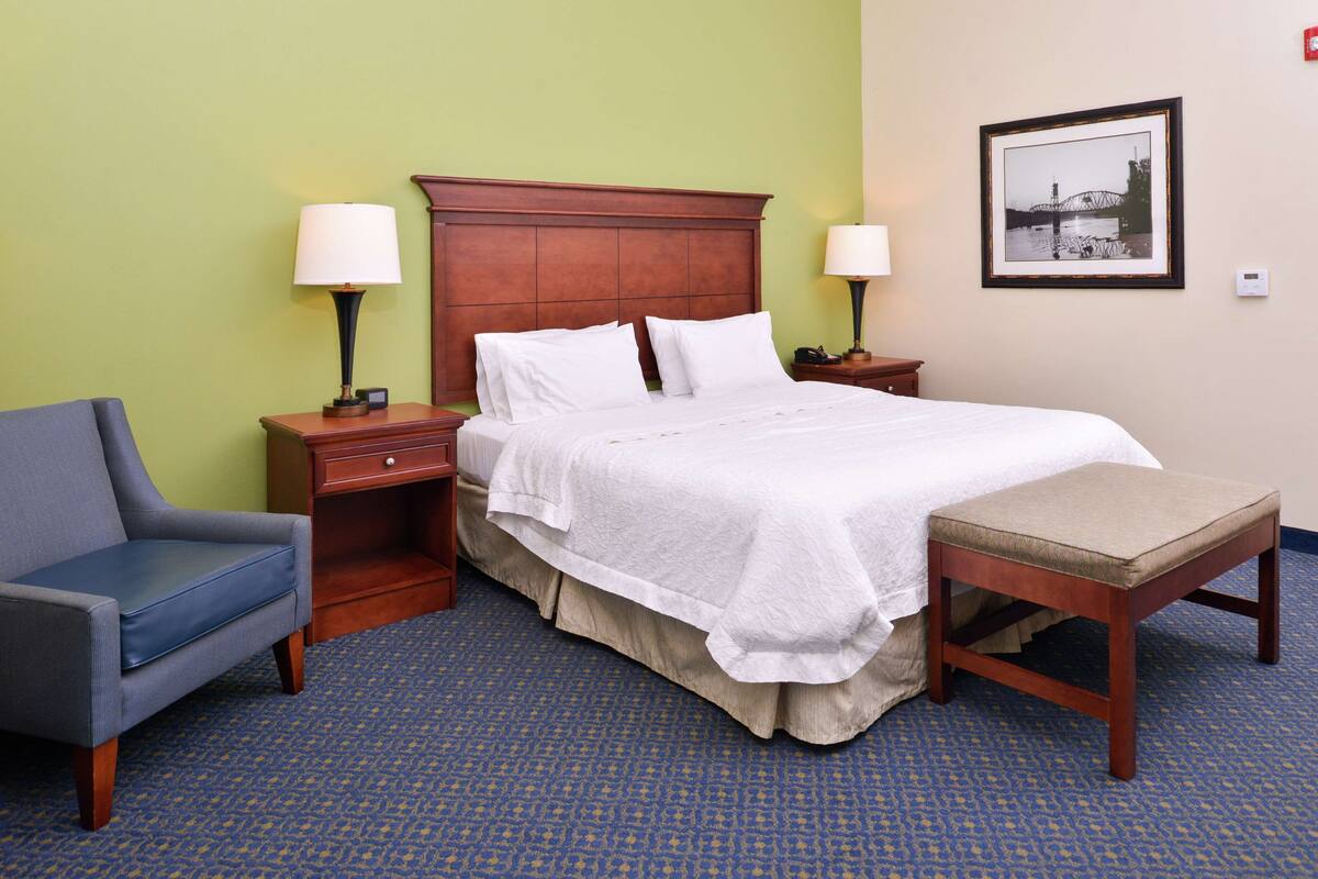 A hotel room with a bed, a chair, a nightstand, a lamp, and a picture on the wall.