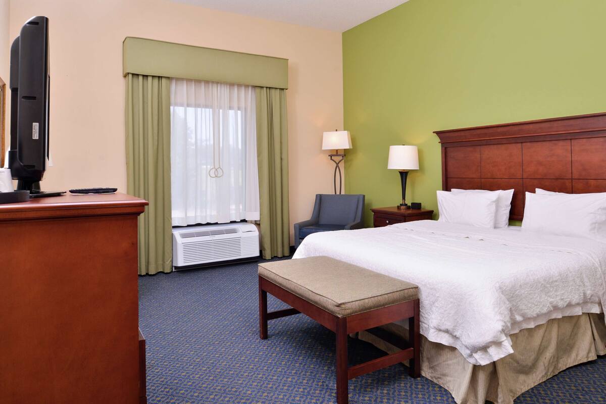 A hotel room with a bed, a desk with a television, a chair, and a lamp.