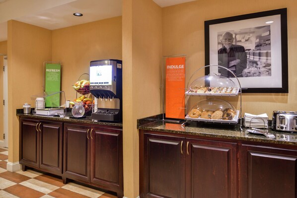 A hotel breakfast area with a coffee machine, fruit bowl, and a display case with pastries.
