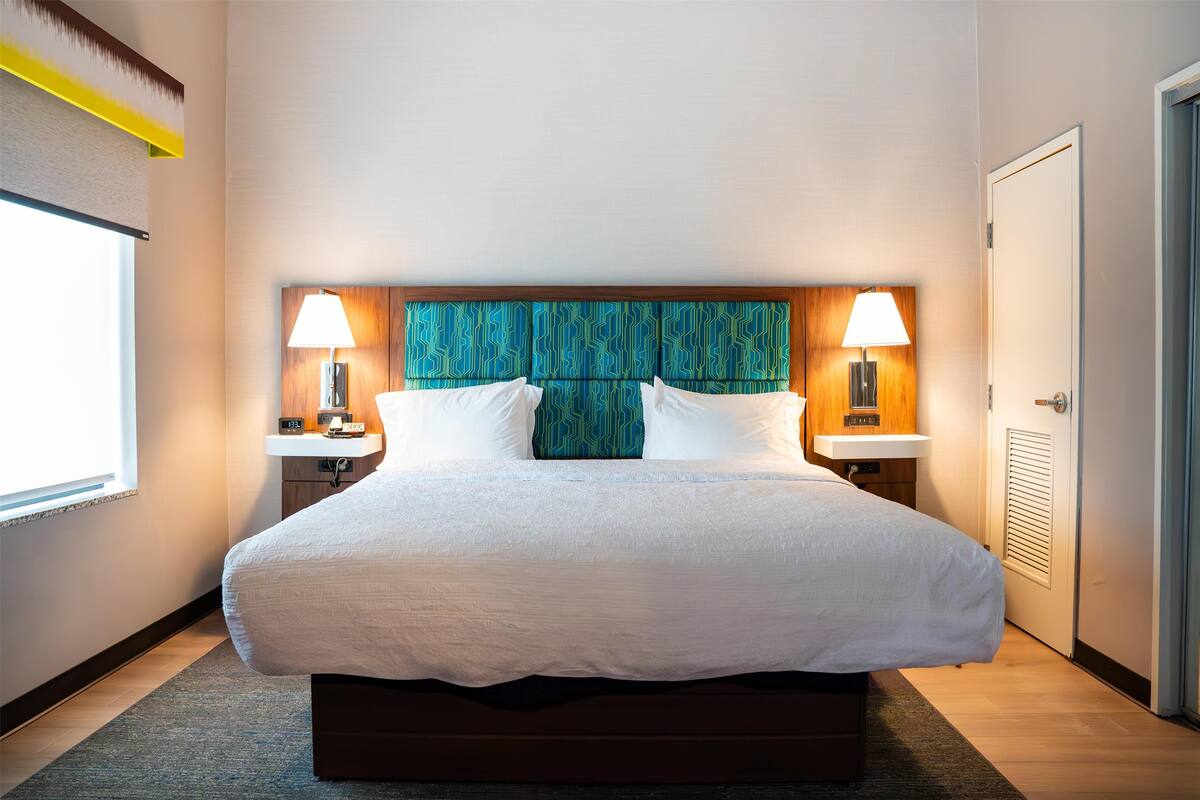 A hotel room with a large bed, two bedside lamps, a wooden headboard, and a window with blinds.