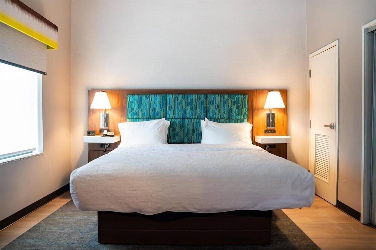 A hotel room with a large bed, two bedside tables, a headboard with a patterned design, and a window providing natural light.