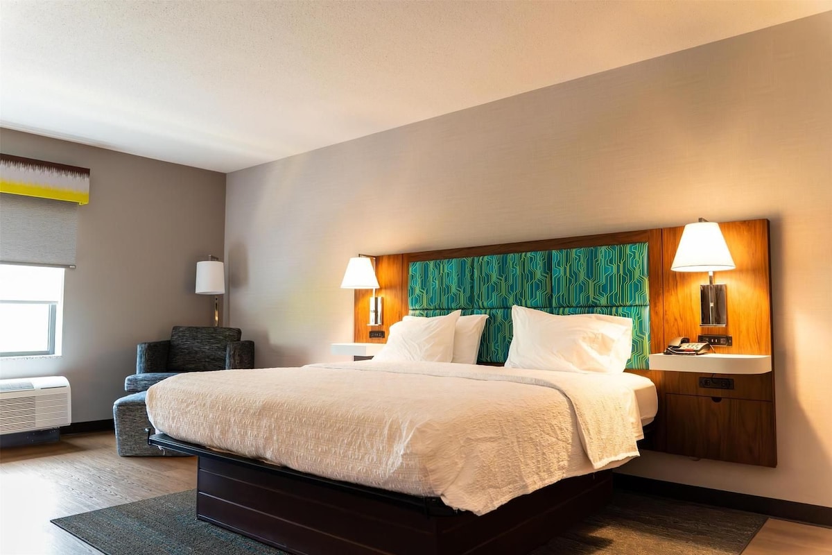 A hotel room with a large bed, two bedside lamps, a headboard with a geometric pattern, a small desk, and a chair.