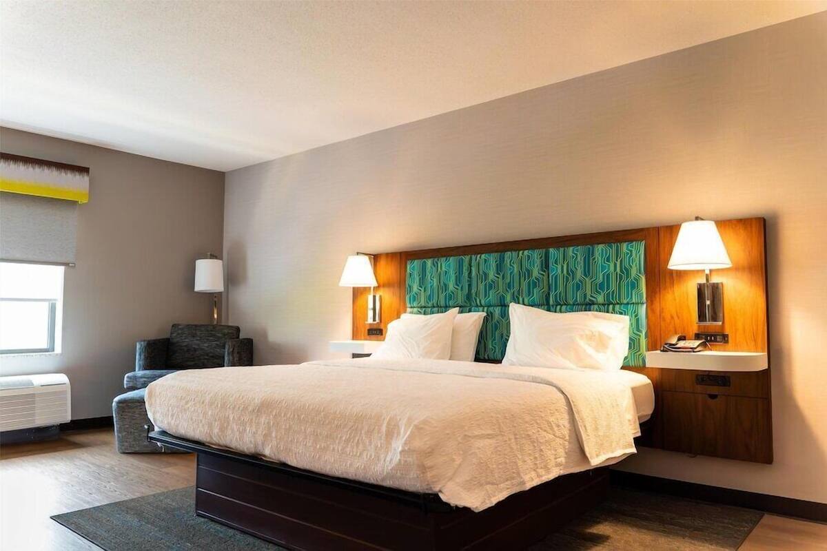 A hotel room with a large bed, two bedside lamps, a small bedside table, a wall-mounted air conditioner, and a window with a view.
