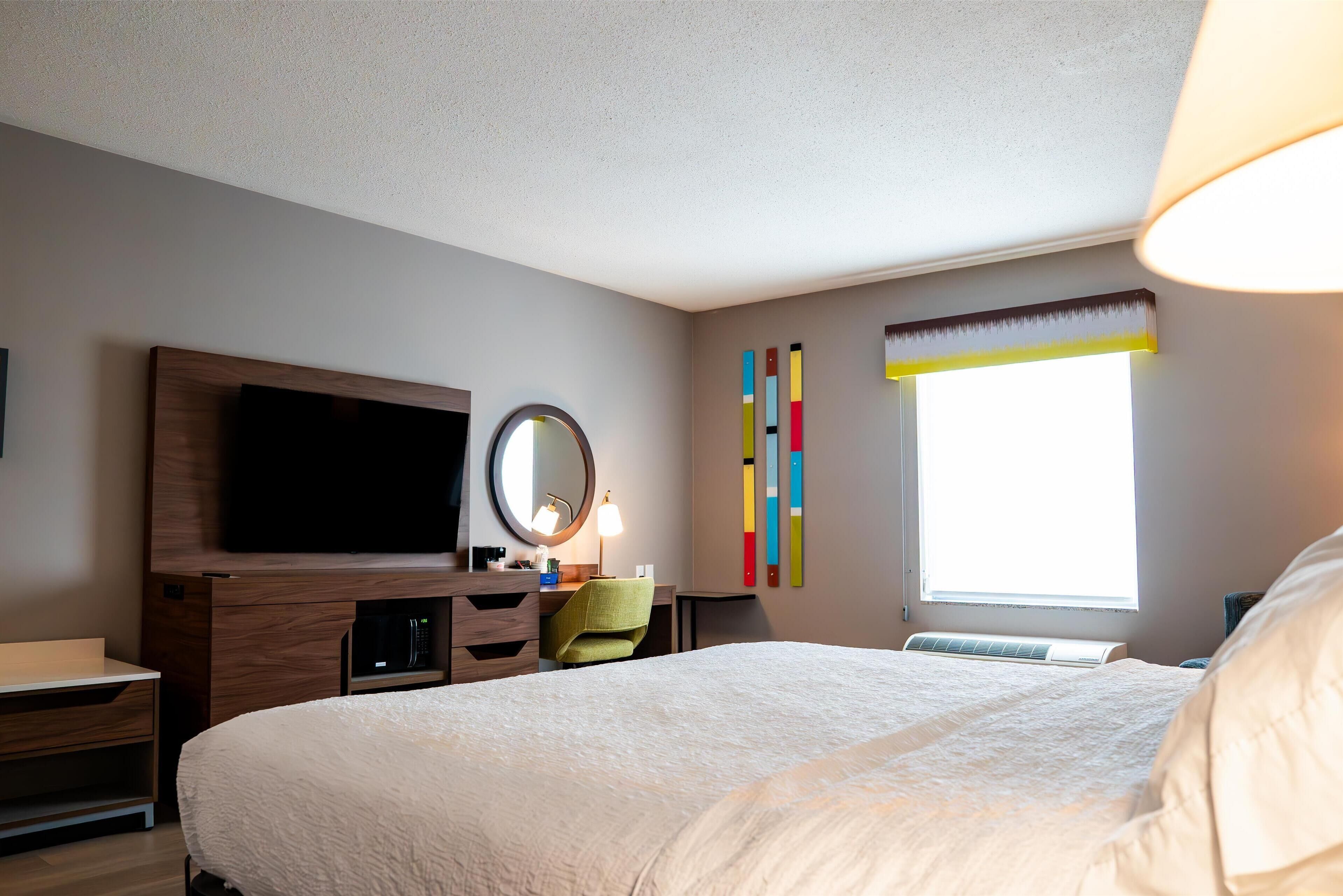 A hotel room with a large bed, a flat-screen TV, a desk with a lamp, a chair, and a window with a view.