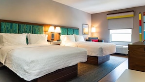 A hotel room with two beds, a nightstand with lamps, a window with a view, and a wall with a colorful artwork.