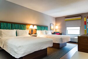 Habitación de hotel con dos camas, una mesita de noche con lámparas, un ventanal con vistas y una pared decorada con una obra de arte colorida.
