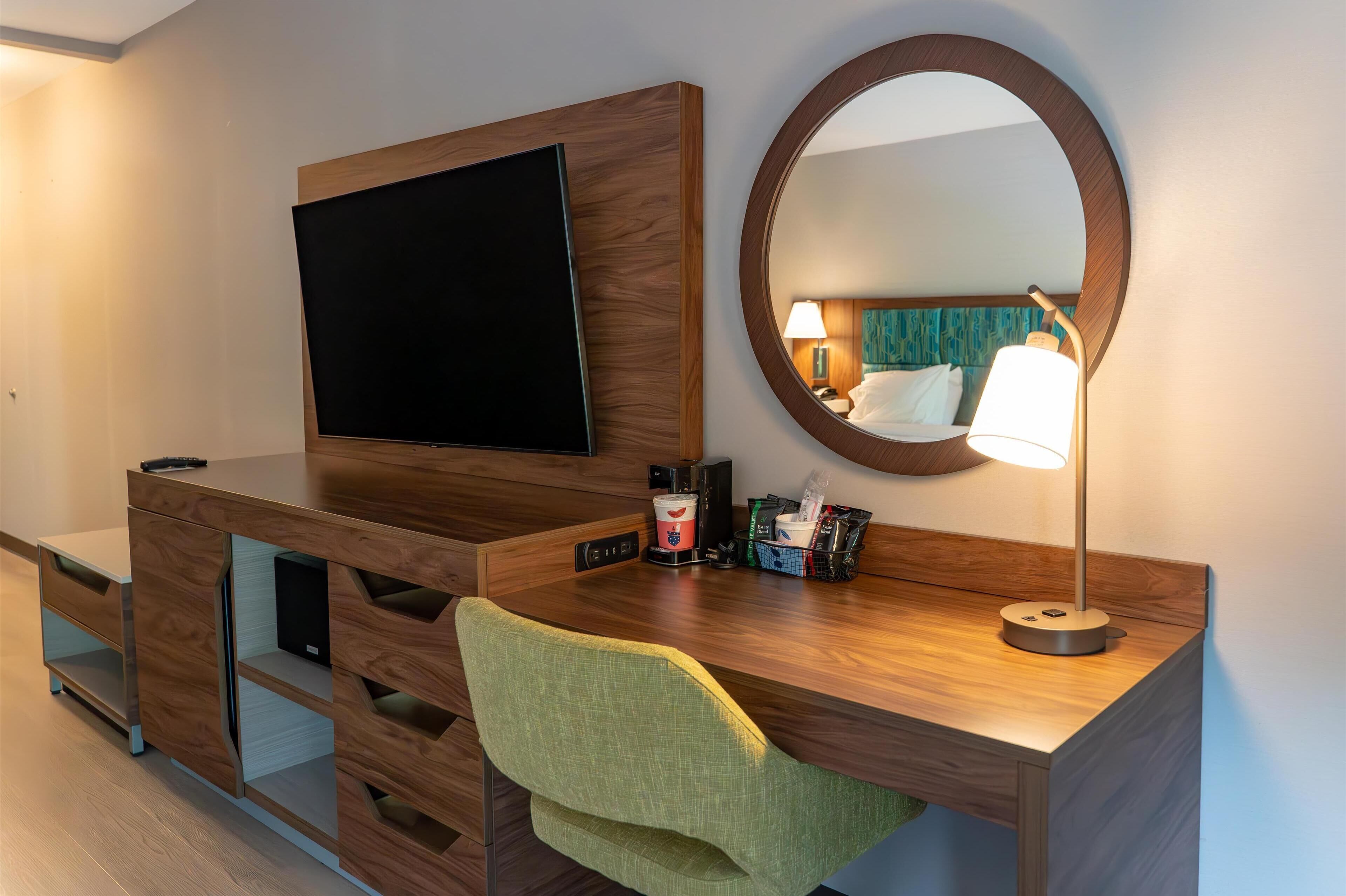 A modern hotel room with a wooden desk, a flat-screen TV, a round mirror, a green chair, and a bedside lamp.