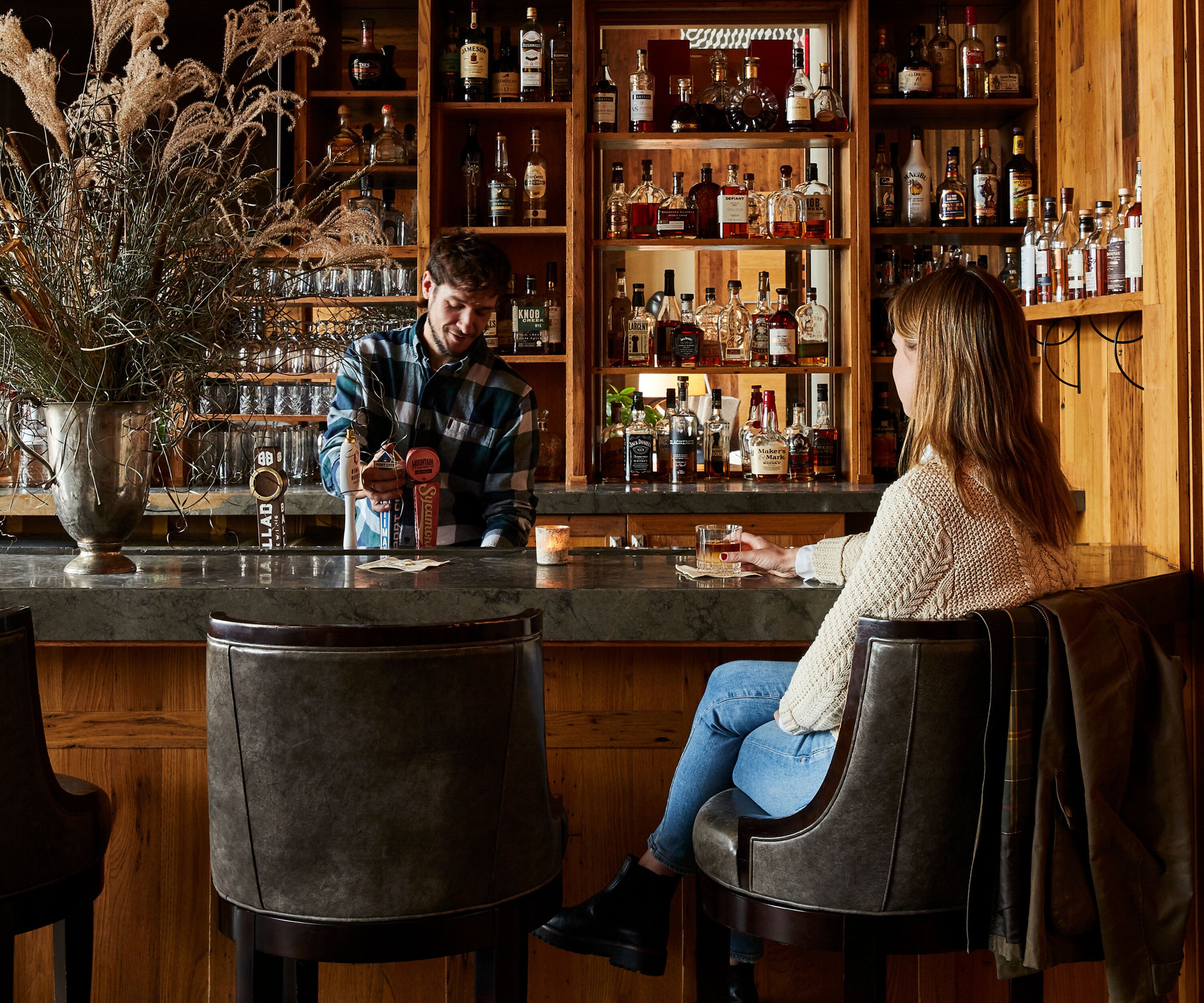 A bar with wooden shelves stocked with various liquor bottles, a bar counter, and two bar stools.