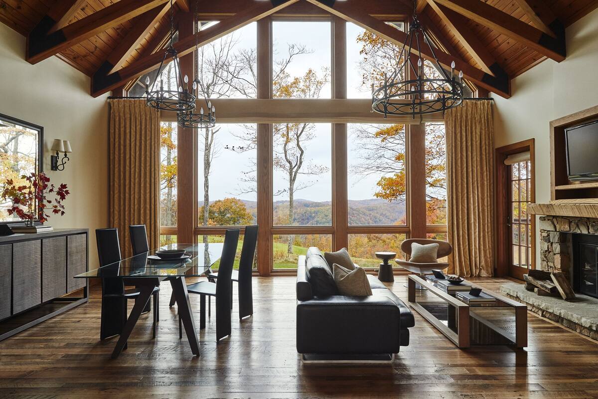 A spacious living room with a large fireplace, a dining area with a glass table, and a view of autumn trees through large windows.