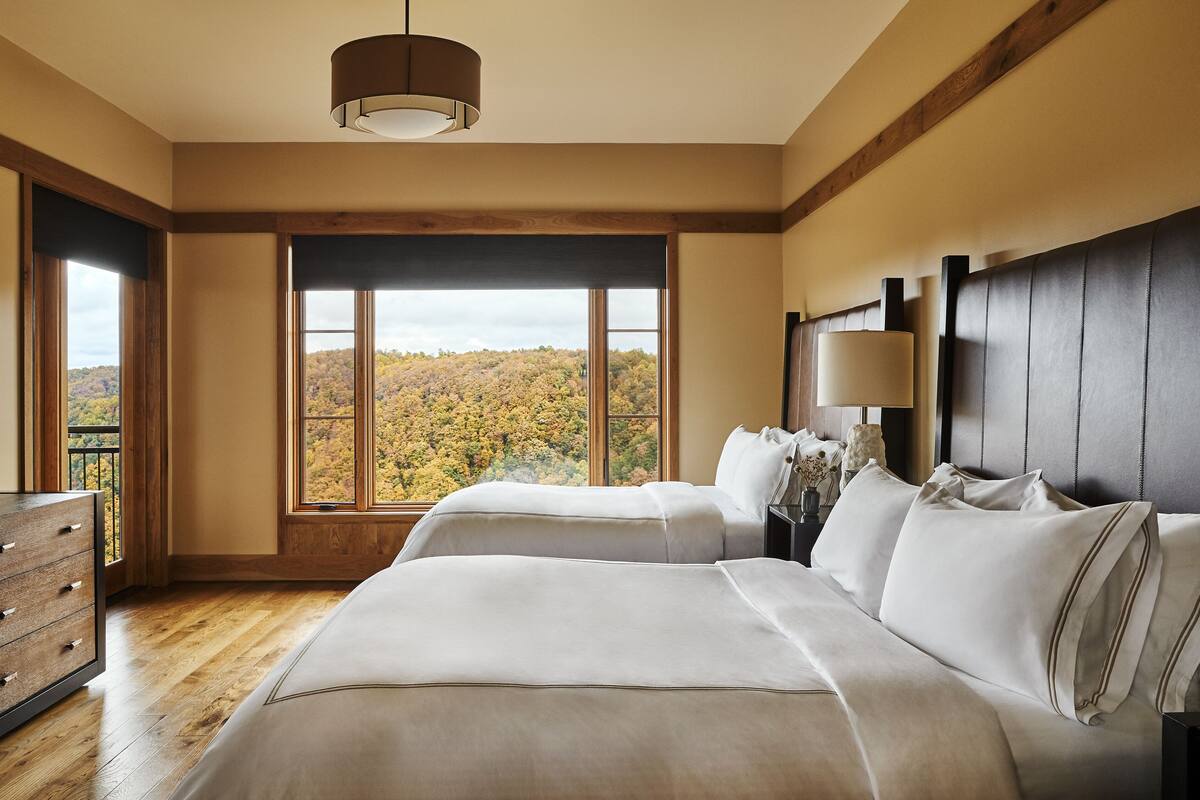 A bedroom with two beds, a dresser, and a large window offering a view of autumn foliage.